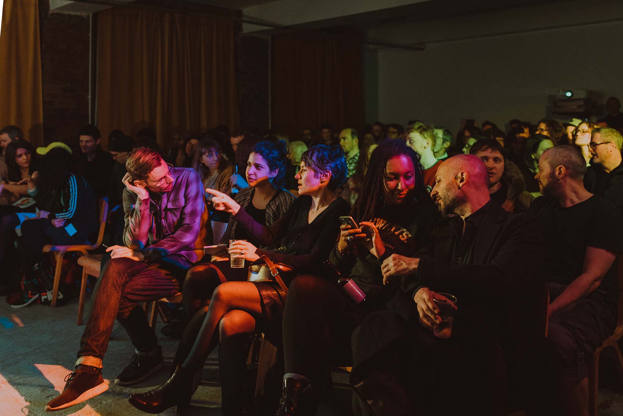 Photo of the award winners at the Glasgow Short Film Festival afterparty at Civic House in 2019. The venue space is full of people sitting in rows on wooden chairs. The room is awash with multicoloured lights in green, pink, red, and blue.