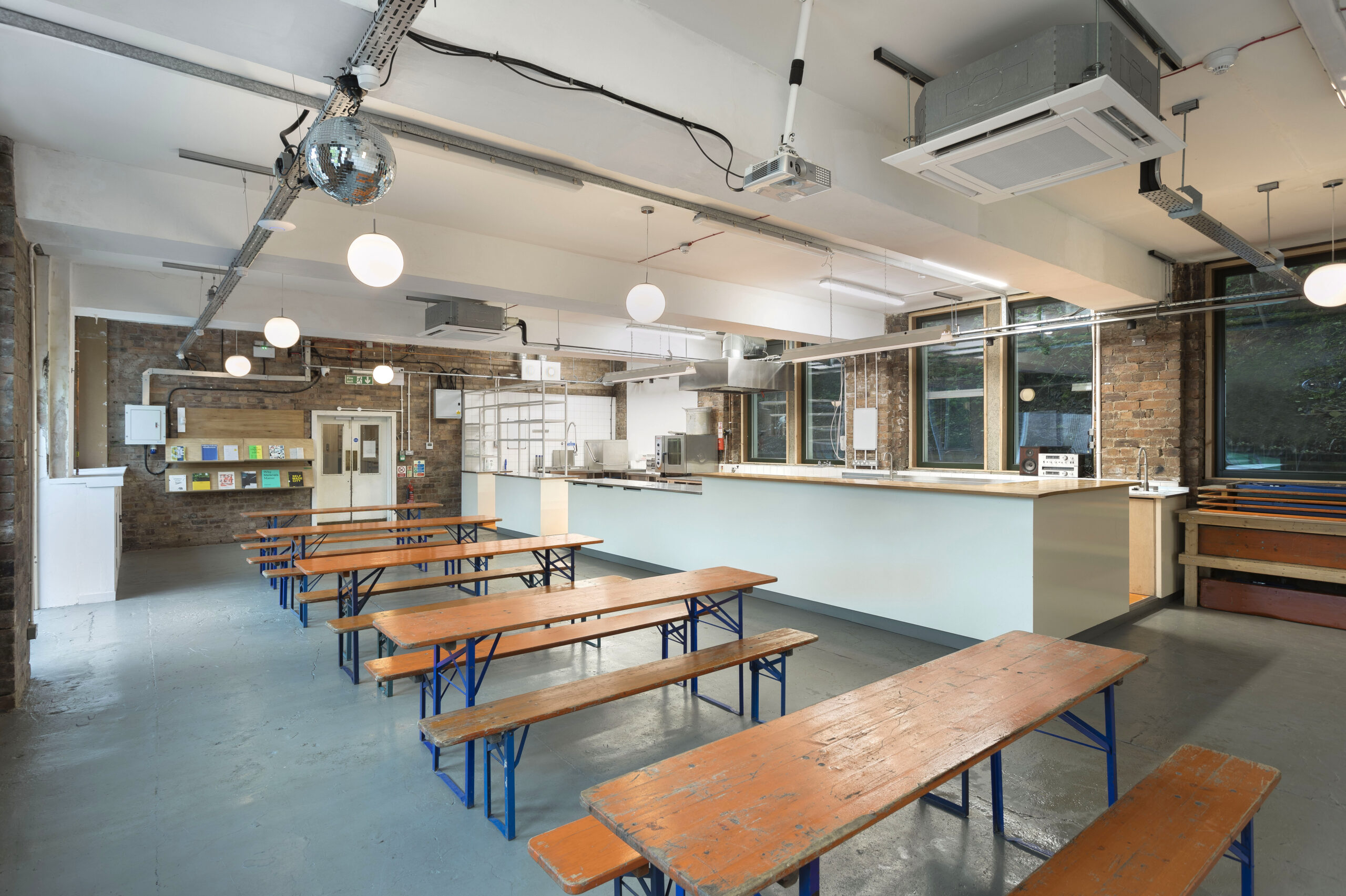 Photo of the Canteen at Civic House. Five orange tables with matching wooden benches stretch down the room from the double doors on the far side. Soft globe pendants add to the light coming in from the windows on the left of the photo. On the right of the photo is he kitchen where Parveen's make their Pakistani inspired dishes. A disco ball hangs from the ceiling in the top left corner.