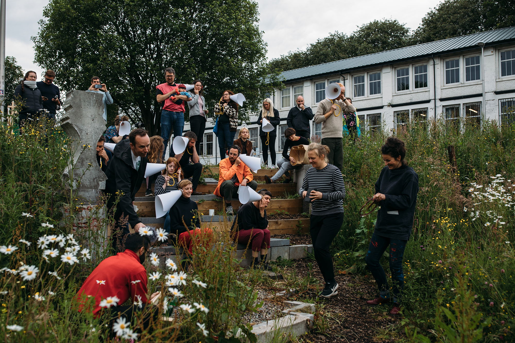 Photo of people gathered in the wildflower meadow outside Civic House for Test Unit 2019. Test Unit 2019 was a summer school for incubating ideas about civic design and making. The participants in this photo hold large cones of paper up to their ears in a listening exercise.