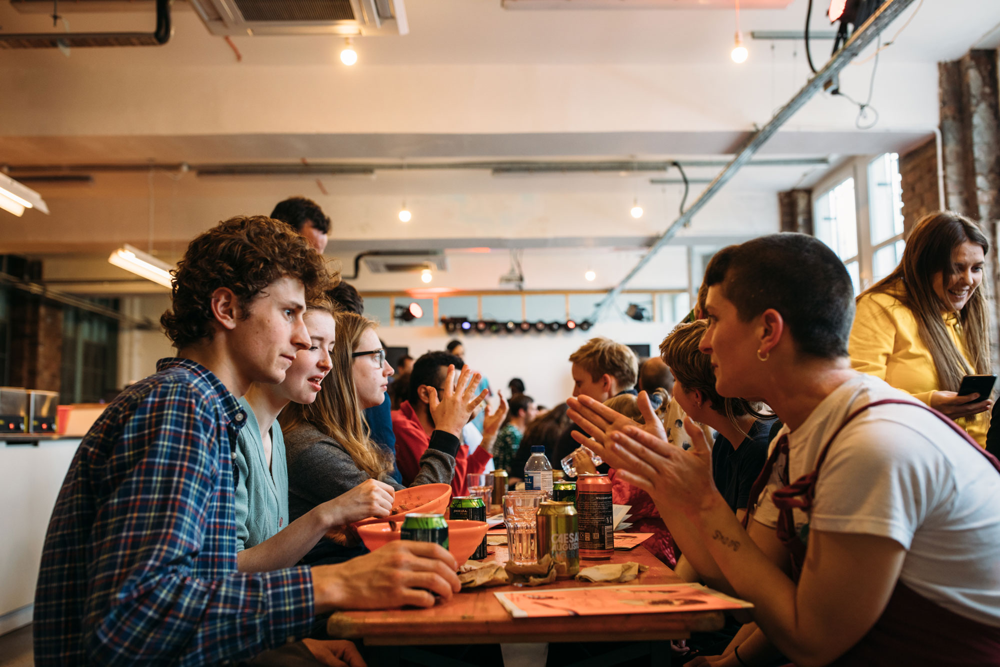 Photo of the Canteen in use at Test Unit 2019. Orange tables are arranged down the length of the room and people engage in conversation over the iconic orange bowls from Parveen's. The space is lit by soft festoon lights and the camera is placed at the head of the table looking along it.