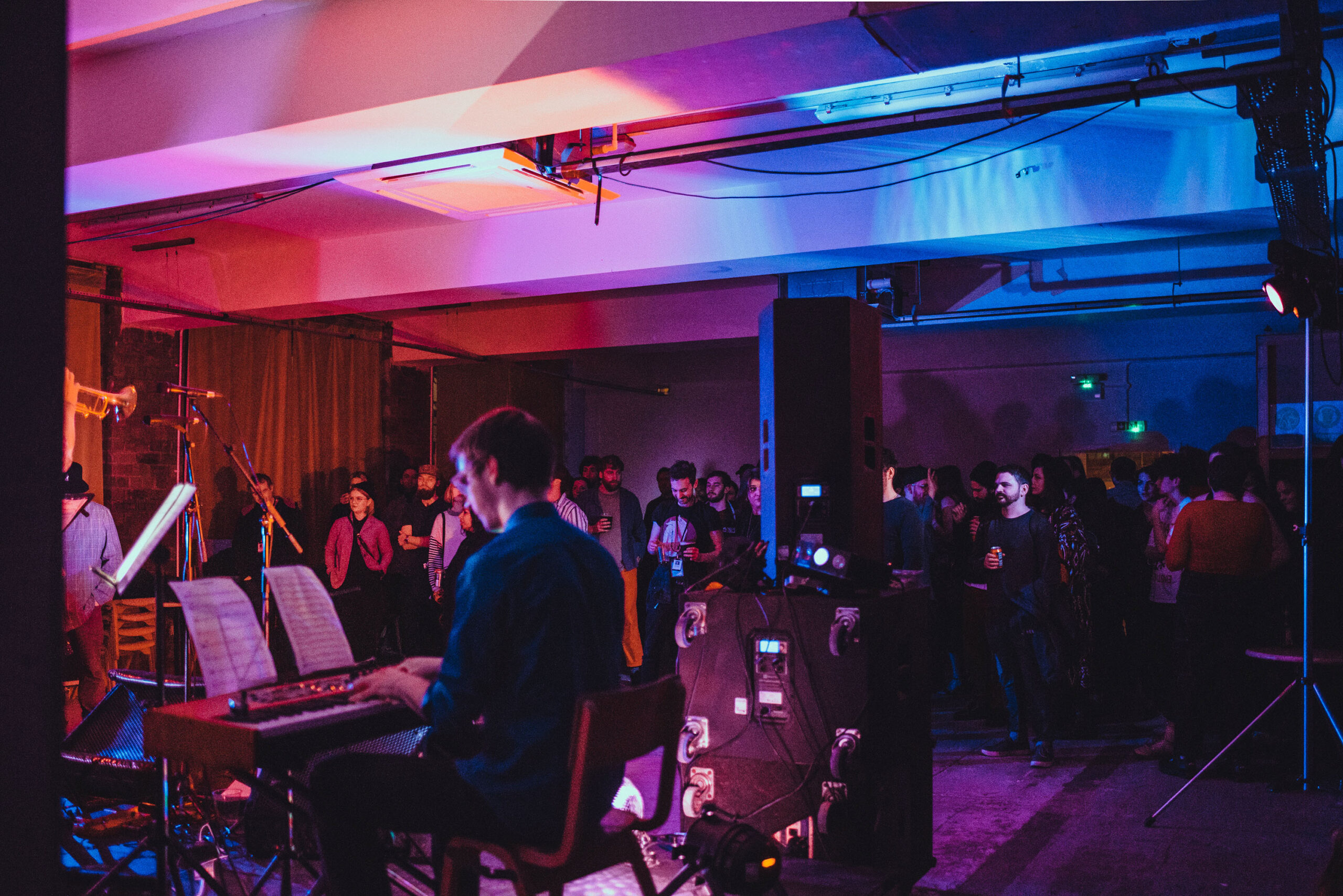 Photo of the afterparty at the Glasgow Short Film Festival 2019 when the ground floor was used as a Festival Hub. The photo shows the Venue space with a piano player in the foreground surrounded by party goers. The venue is bathed in pink, red, and blue lights. The end of a trumpet can just be recognised on the left of the photo.