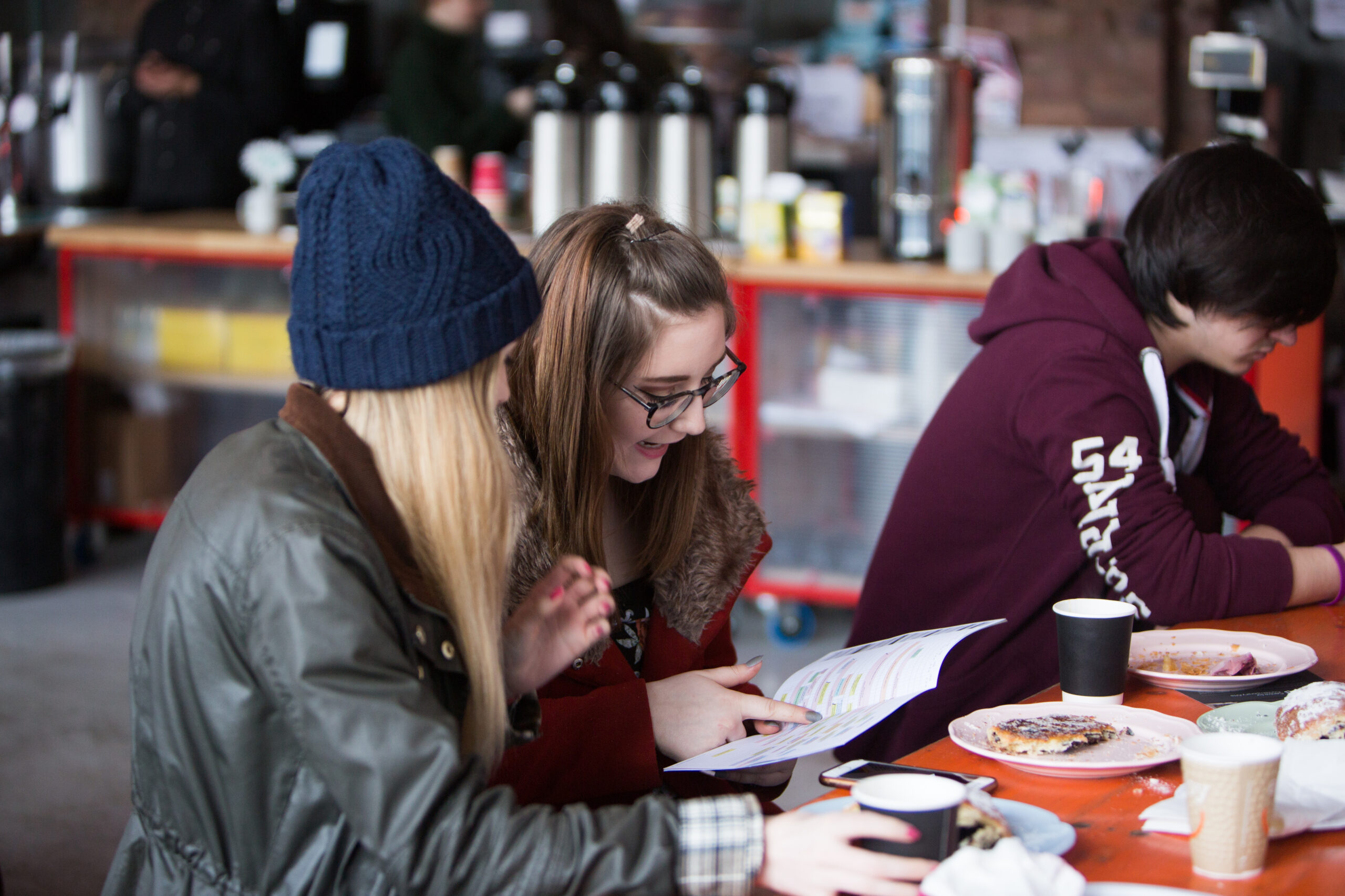 Photo of attendees at the Glasgow Short Film Festival 2018. Three attendees are sat in the canteen which is being used as part of the Festival Hub. Two of the attendees are pointing out something of interest in the program while snacking on cakes and coffee at one of the iconic orange tables. Coffee urns line the background of the shot.