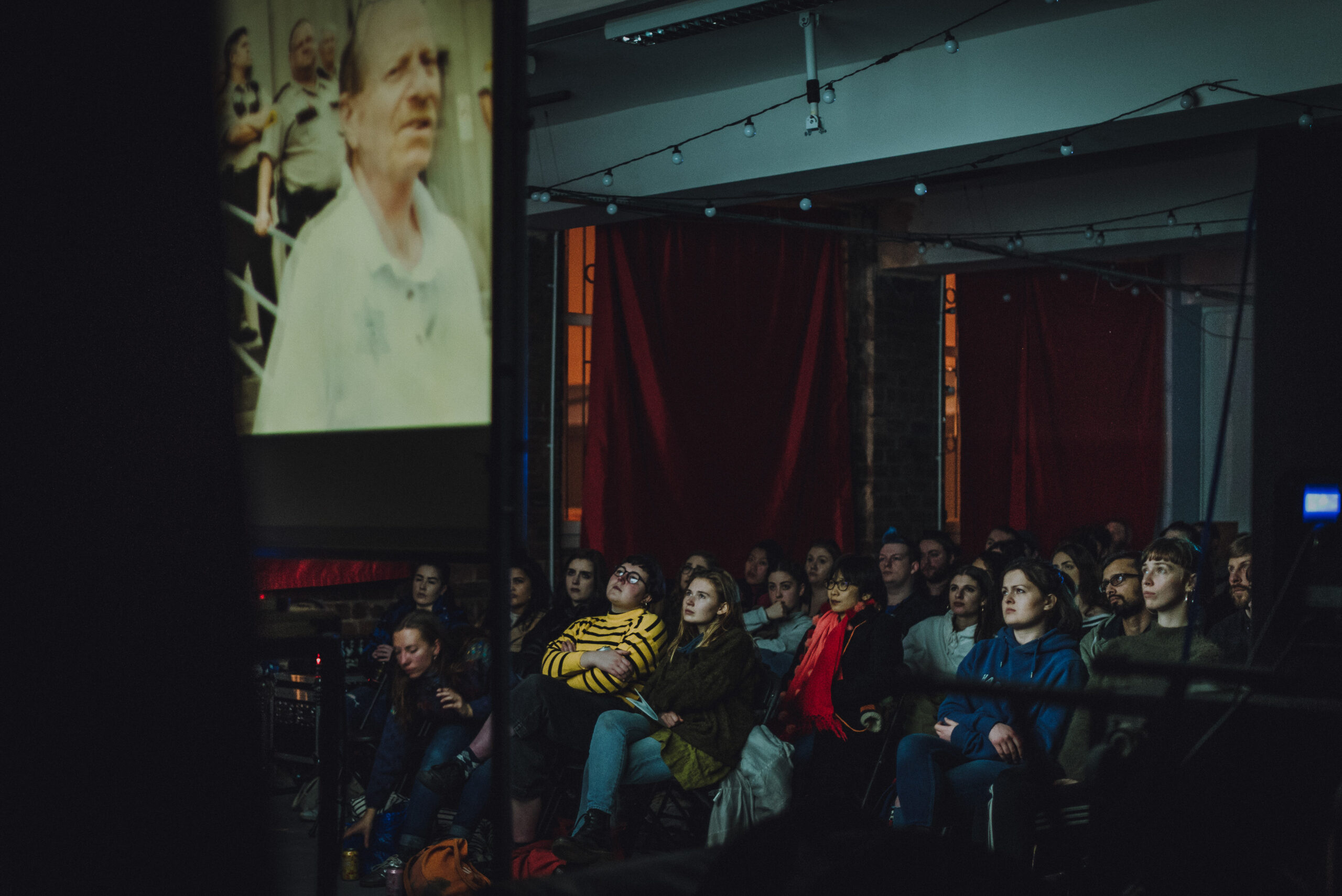 Photo from Glasgow Short Film Festival 2022 when Civic House was booked as a Festival Hub. The photo shows a darkened venue space illuminated by a projection of a film onto a screen in the foreground. The faces of the audience are illuminated by the projection in the foreground.