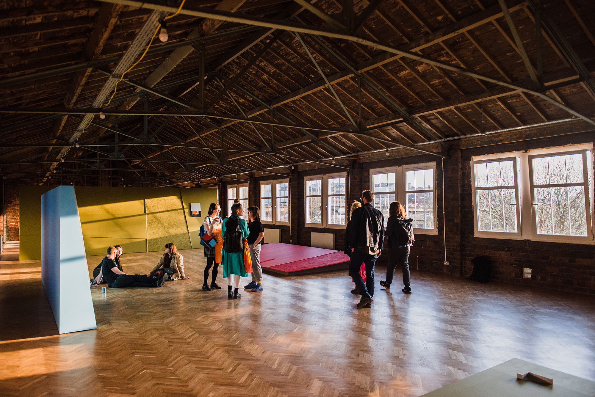 Photo of the coworking space as it was set up for the workshop by Toby Paterson. Three large geometric structures are placed playfully about the industrial space. They are blue, yellow, and red. A group of people are conversing and sitting on the floor as light comes in from windows to the right of the photo.