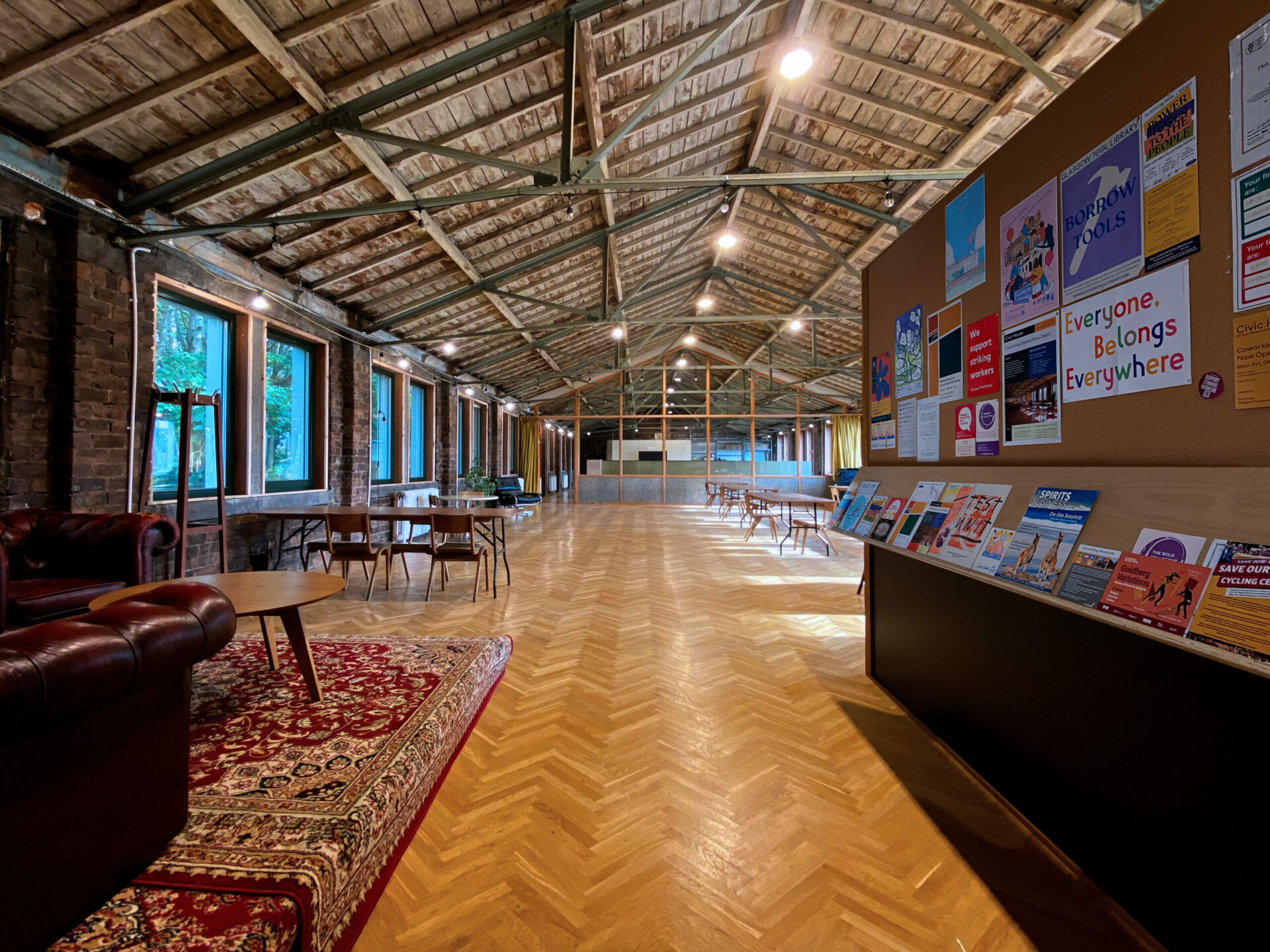 Photo of the first floor coworking office. Red couches can be seen on the left and various posters about events in the space and coworking members can be seen on the notice board on the right. The length of the hall is hung with festoon lighting, and light comes in from windows on both sides.