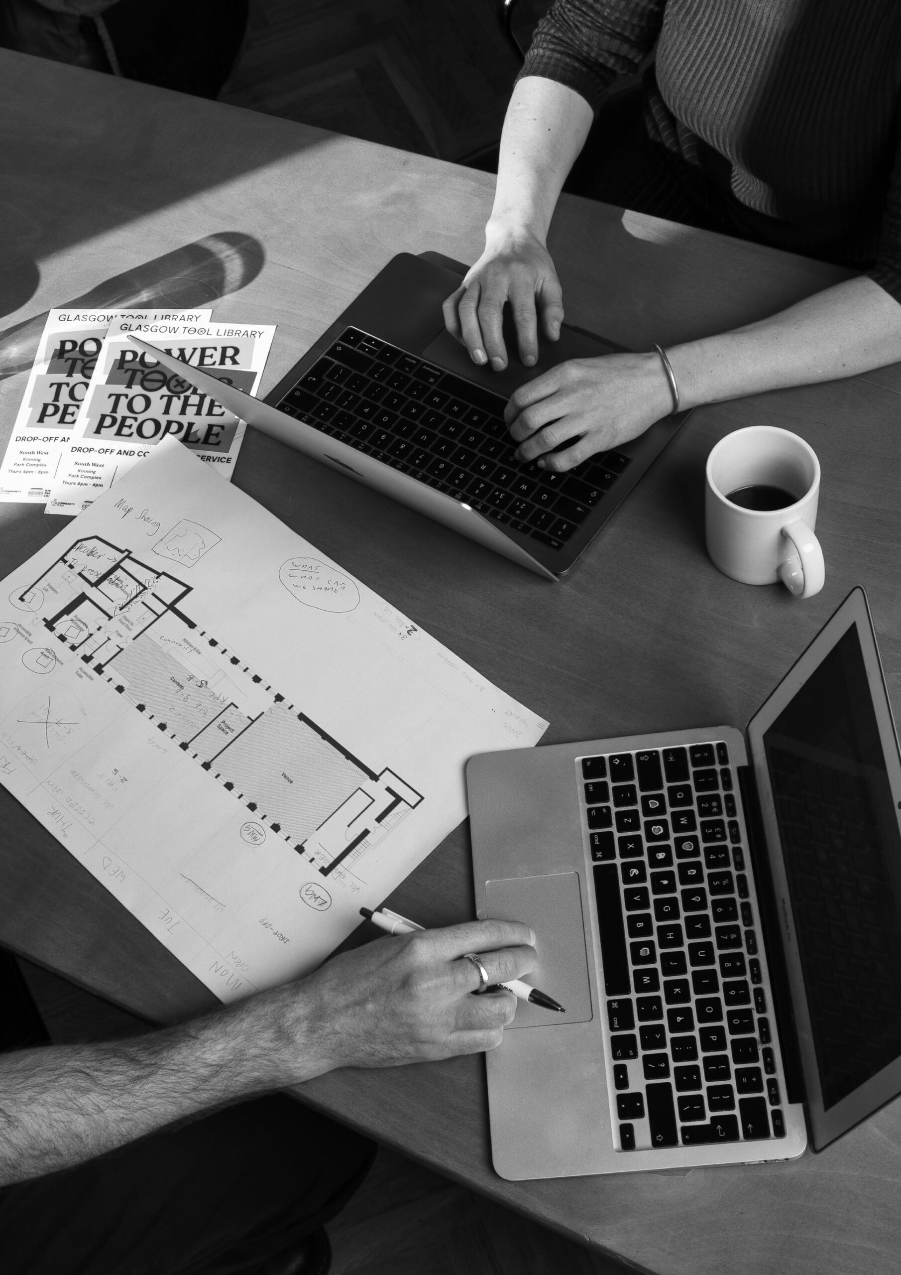 A black and white photo illustrating collaborative coworking at Civic House. An annotated floor plan sits at an angle between open laptops, cups of coffee, and flyers for Glasgow Tool Library.