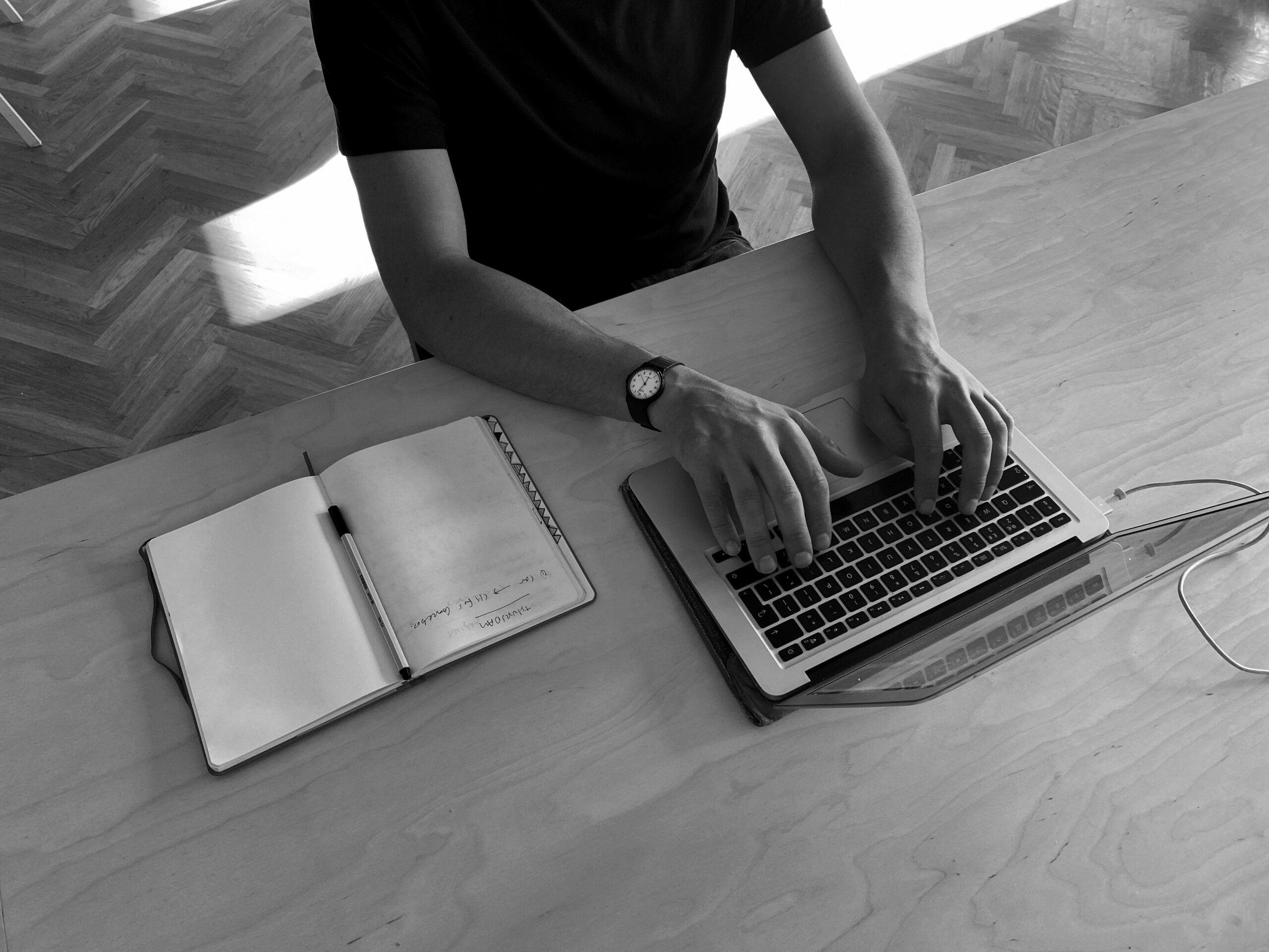 A black and white photo of a hot coworking member typing at a MacBook with an open notebook to their right. You can see the parquet flooring below.