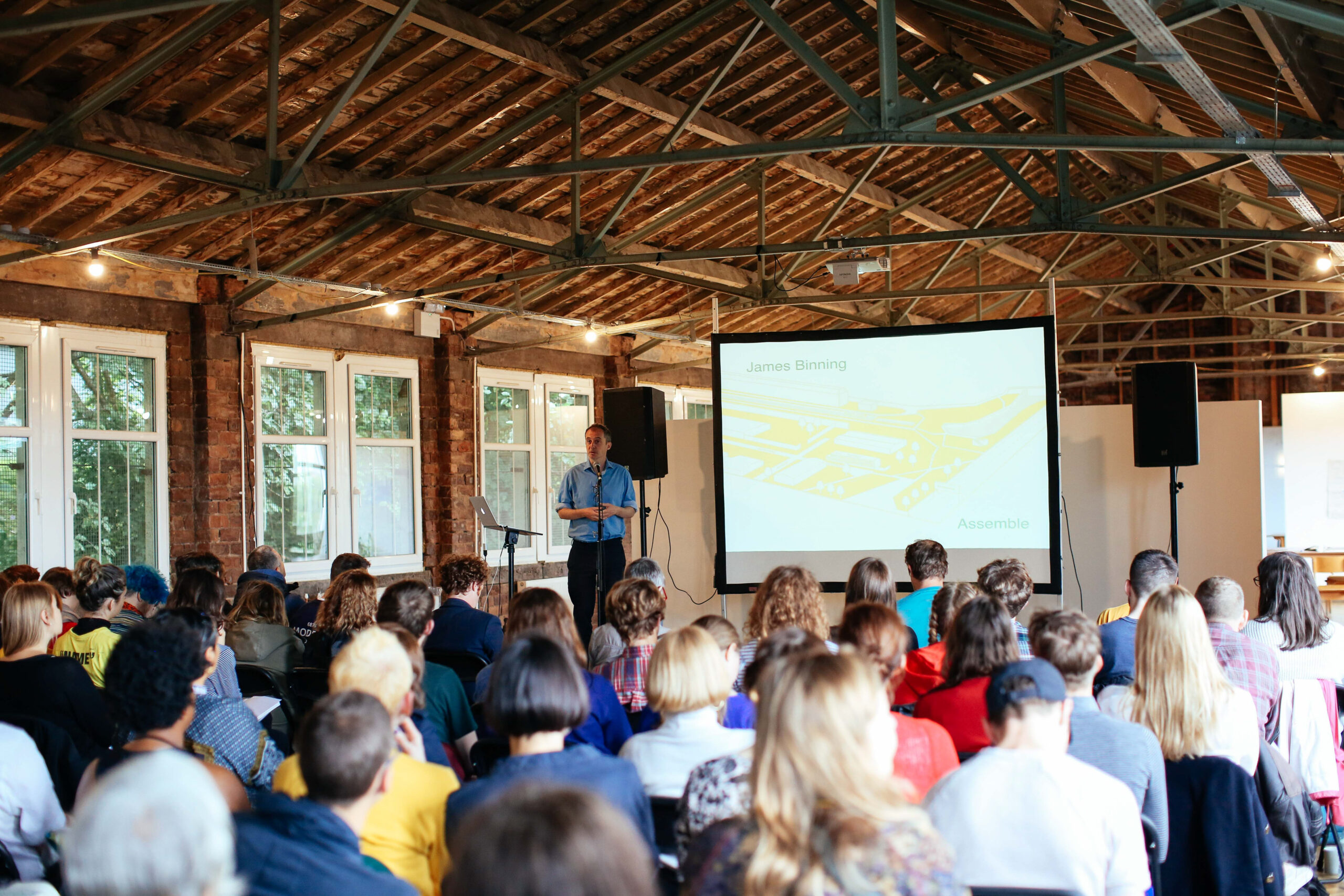 A photo of the Assemble x Test Unit event in 2018. A man stands at the back of the picture presenting with a microphone, laptop on a stand, projector screen, and speakers. In the foreground is a rapt audience. Light comes in from the windows and you can see festoon lights against the beams of the ceiling.