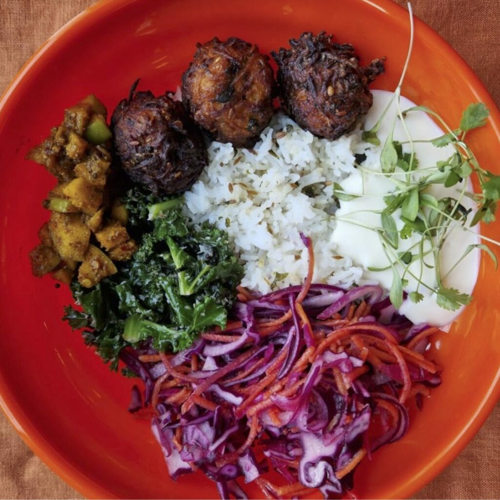 Photo of a lunch bowl by Parveen's the Canteen here at Civic House. It is an example of the lunch that comes with the Civic House Studio Day Pass.The bowl is one of the iconic plastic, orange bowls that can be found in the Canteen or upstairs in the coworking space. Inside the bowl is rice, raita, pickled onion, falafel, kale, and marinated tofu.