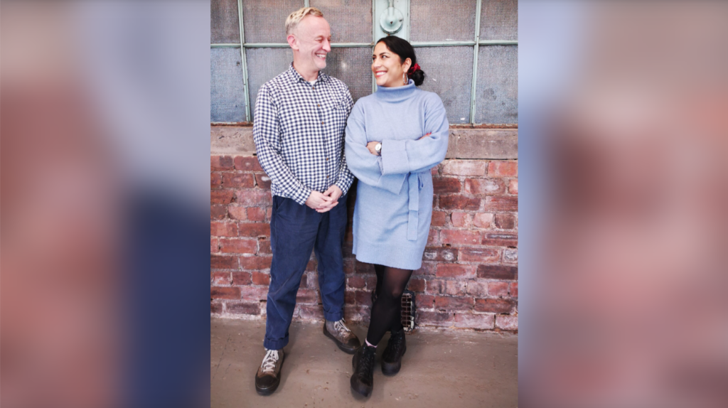 Photo of Paul Crawford and Prerna Menon, coworking members who founded Panther Milk. The photo is taken in the venue space against the exposed brickwork and industrial windows. Paul and Prerna stand next to each other with their arms crossed. Paul is wearing a checkered button-down shirt, jeans and a pair of brown shoes. He has short cropped hair and is smiling at Prerna. Prerna is wearing a pstel blue dress with a high collar, tights, and black ankle boots. She is grinning at Paul.