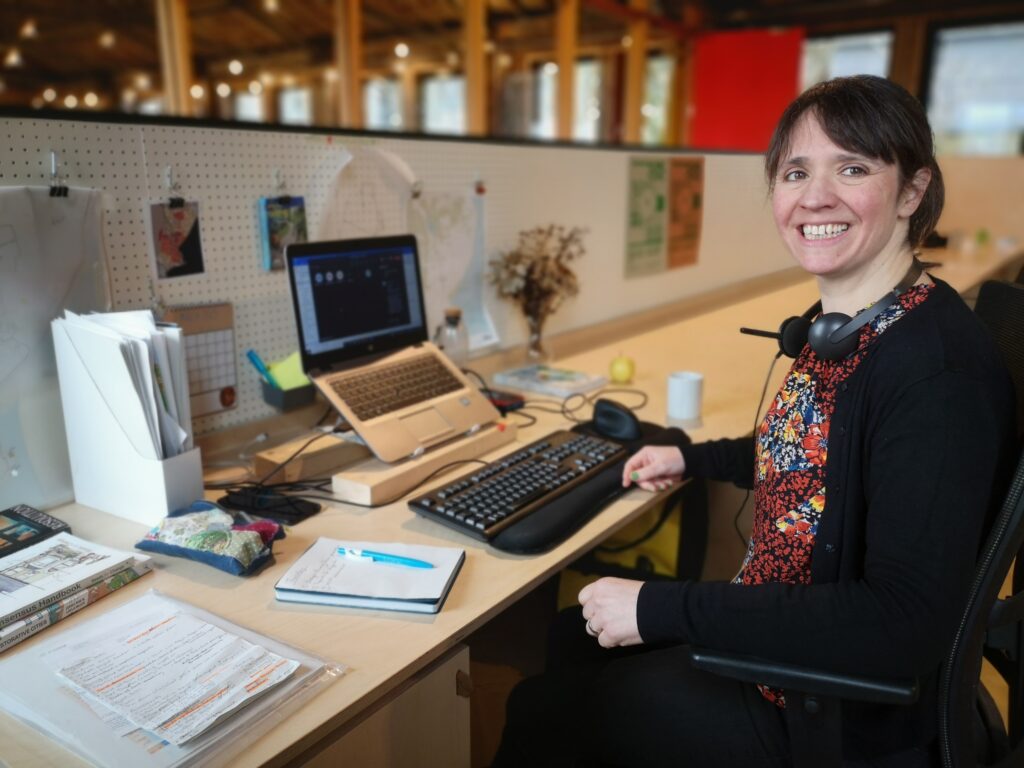 Photo of Alex Wilde, coworking member for the Shaping Places for Wellbeing programme. Alex is sitting in the Fixed Desk area of the coworking studio on the first floor. Her laptop is set up with a stand, keyboard, and mouse and is surrounded by notebooks and paper trays. It is an organised and busy workspace. Alex is wearing a black cardigan over a floral shirt with a pair of headphones about her neck. She is turned towards the camera and is grinning.