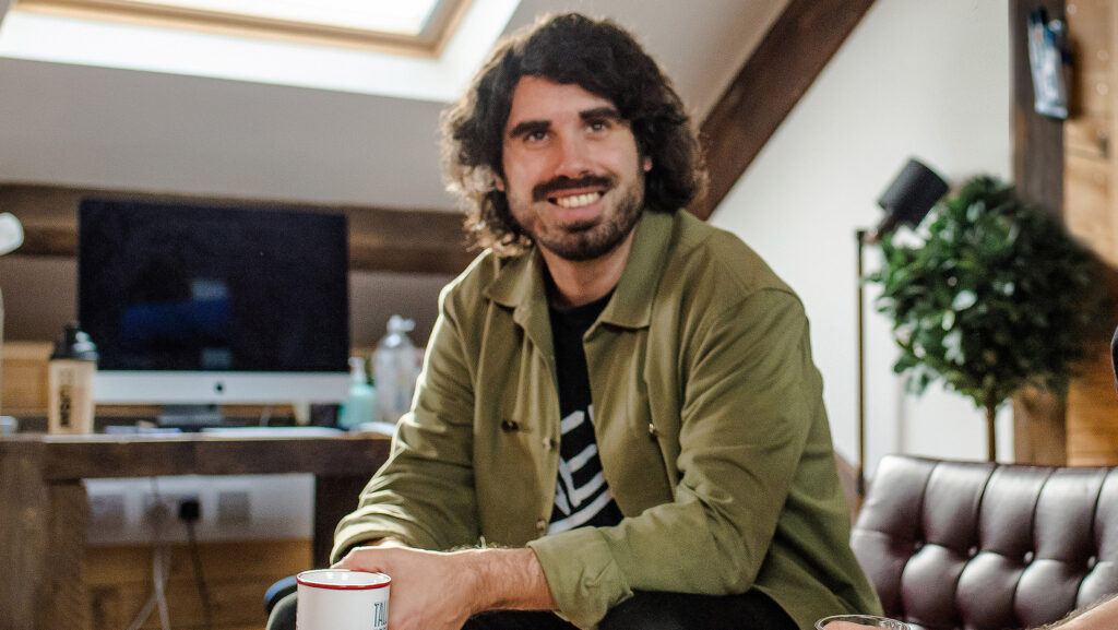 Photo of Mark Martin, coworking member with Rabbithole, a brand identity web studio. The photo depicts mark in a professional space with wooden beams and a midcentury Barcelona leather chair. An Apple Mac is visible in the background alongside an indoor plant and a lamp. Mark is sitting in the foreground wearing an olive green shirt over a black graphic t-shirt. He has mid-length hair and a cropped beard and is smiling at the camera.