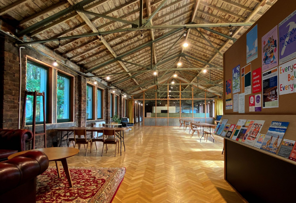 Photo of the first floor coworking space. Red couches can be seen on the left and various posters about events in the space and coworking members can be seen on the notice board on the right. The length of the hall is hung with festoon lighting, and light comes in from windows on both sides.