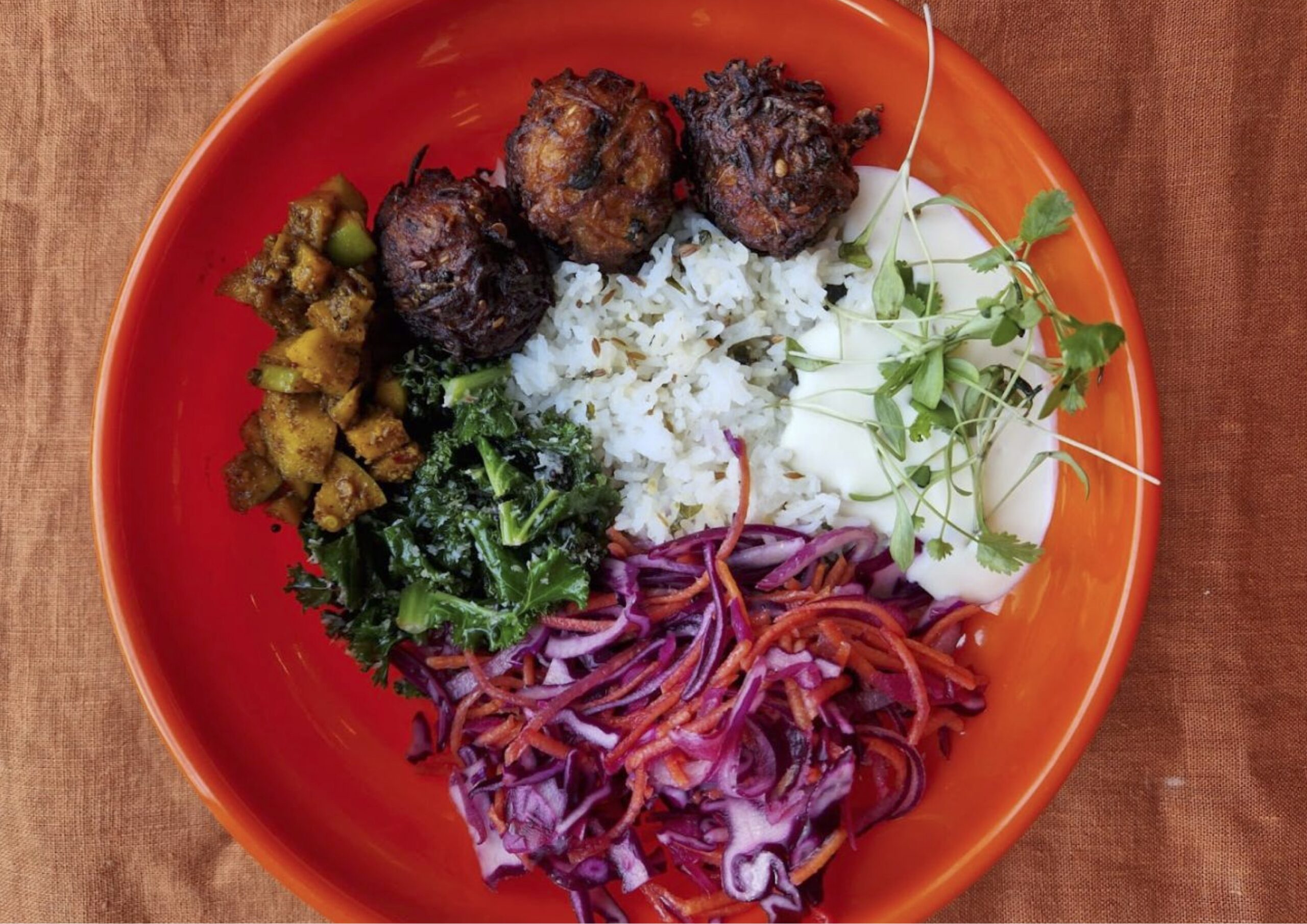 Photo of a lunch bowl by Parveen's the Canteen here at Civic House. It is an example of the lunch that comes with the Civic House Studio Day Pass.The bowl is one of the iconic plastic, orange bowls that can be found in the Canteen or upstairs in the coworking space. Inside the bowl is rice, raita, pickled onion, falafel, kale, and marinated tofu.