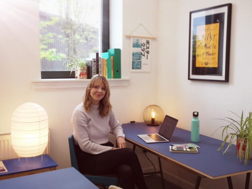 Photo of Jade Sturrock, coworking member with Glasgow Connected Arts Network. Jade is sitting in the ground floor Accessible Workspace. She is surrounded by soft Scandinavian lamps on blue trestle tables. Her laptop is open and is smiling at the camera. Light comes in from a window behind Jade. The room is decorated with framed risographed posters, books, and spider plants. Jade is wearing a soft white, ribbed jumper.