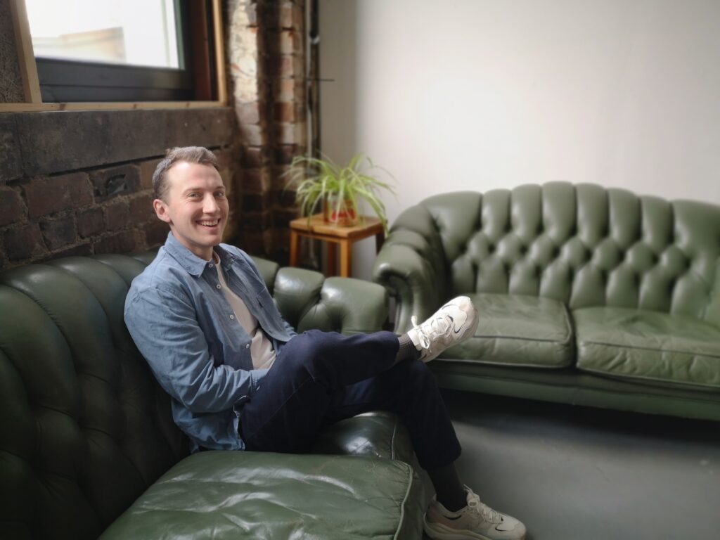 Photo of James Bryson, coworking member with Placeshapers, the national network of place-based housing organisations. James is siting on one of two green chesterfield-style couches in the Venue Space on the ground floor. He is surrounded by exposed brickwork and spider plants. James is wearing denim shirtsleeves over a white t-shirt, navy trousers and white trainers. His hair is shirt and he is smiling at the camera with one leg crossed over the other.