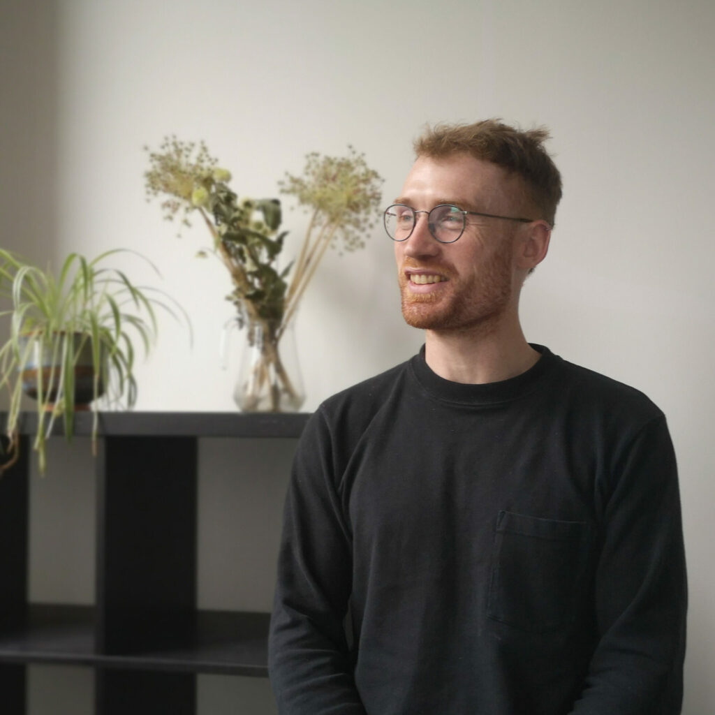 Photo of Graham McGrath, coworking member with Local Energy Scotland. Graham is sat in the Project Room with natural lighting and plants in the background. He is dressed in a dark grey shirt with a breast pocket. He wears round rim glasses with a cropped beard and short ginger hair. He is smiling and looking off to the left of the photo.