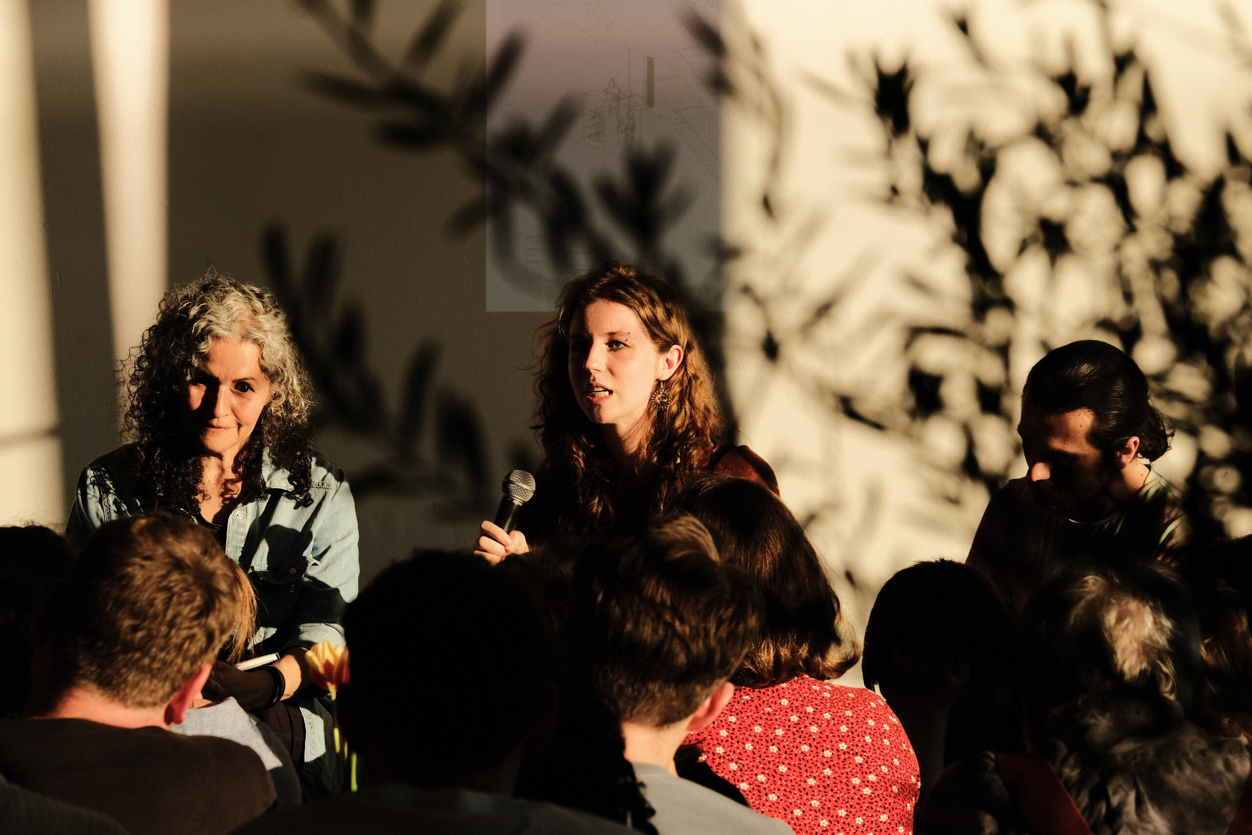 Photo of the Festival Hub as it was used as part of the Architecture Fringe 2023. Three panel members sit in front of an architectural projection. There is the shadow of trees outside cast across the image. The foreground is crowded with a rapt audience.