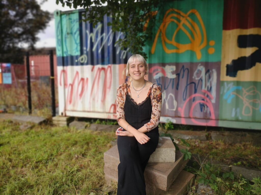 Photo of social change hot desk user Leah Cowling. Leah is sat outside on some cement blocks with artfully gratified shipping containers behind her. She is smiling at the camera and has a platinum blonde bob.