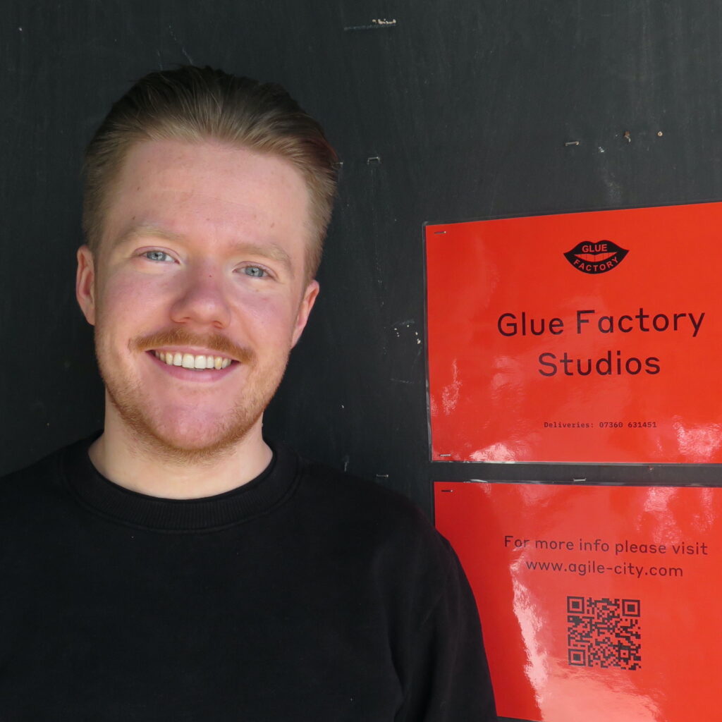 Photo portrait of the Glue Factory's new studio and production space manager Fraser Whiting. Fraser is standing in front of a black wall. Next to him is a red sign that says "Glue Factory Studios." Fraser is wearing black and is smiling at the camera. His hair is blonde and slicked back and he has a moustache.