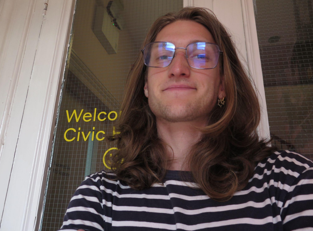 Photo of Sebastian Taylor the Front of House and Communications Assistant in the Coworking Studio at Civic House. Sebastian is standing in front of the entryway to Civic House, and the words "Welcome to Civic House" can be seen in linoleum on the window behind them. They have shoulder length blonde hair, glasses, and are smiling at the camera.