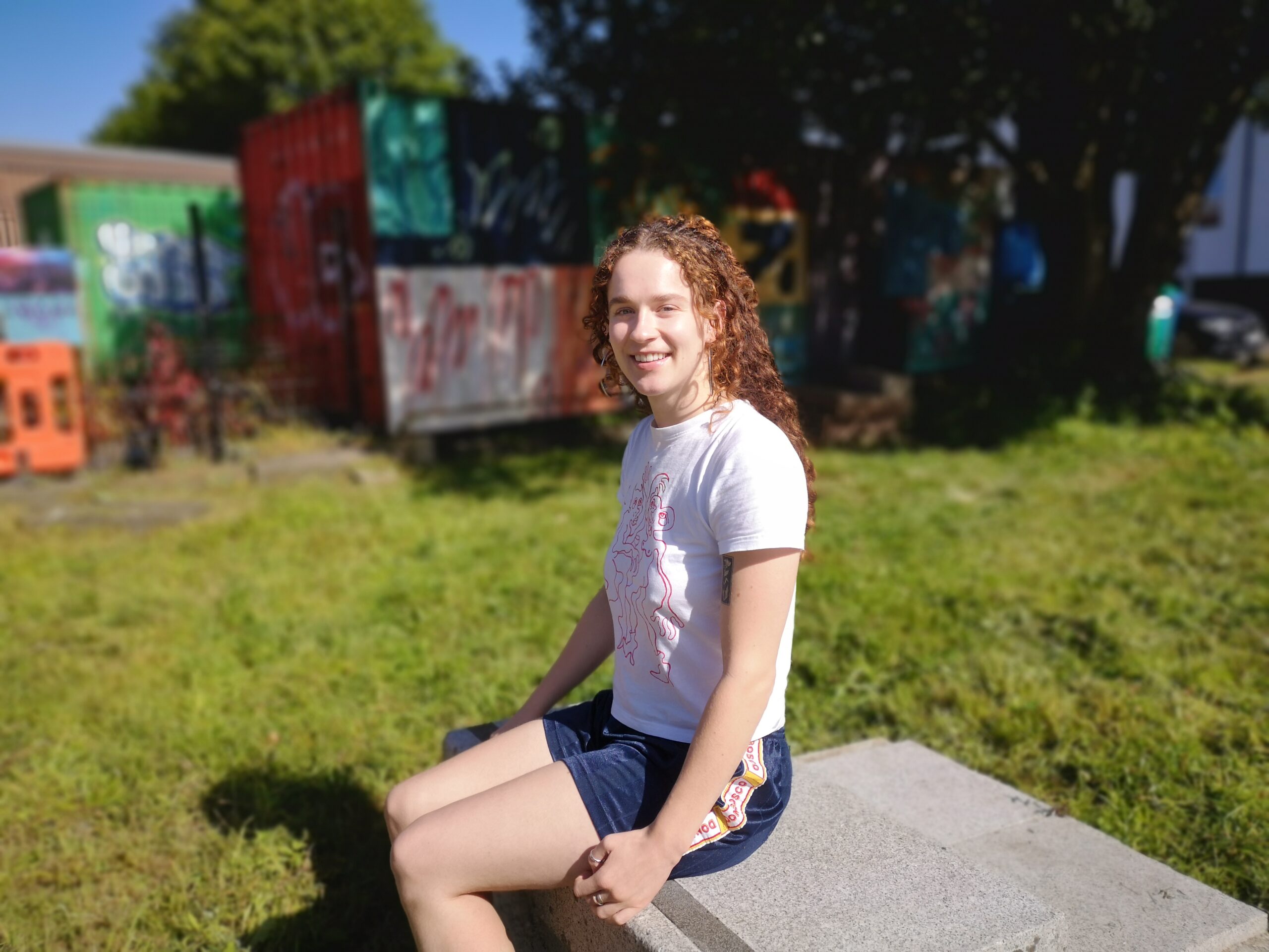 Photo of Hot Desk Member for Ethical Practice Sorcha Perris. Sorcha is sitting outside on a cement block surrounded by green grass. It is sunny and she is in shorts and a t-shirt and is smiling at the camera.