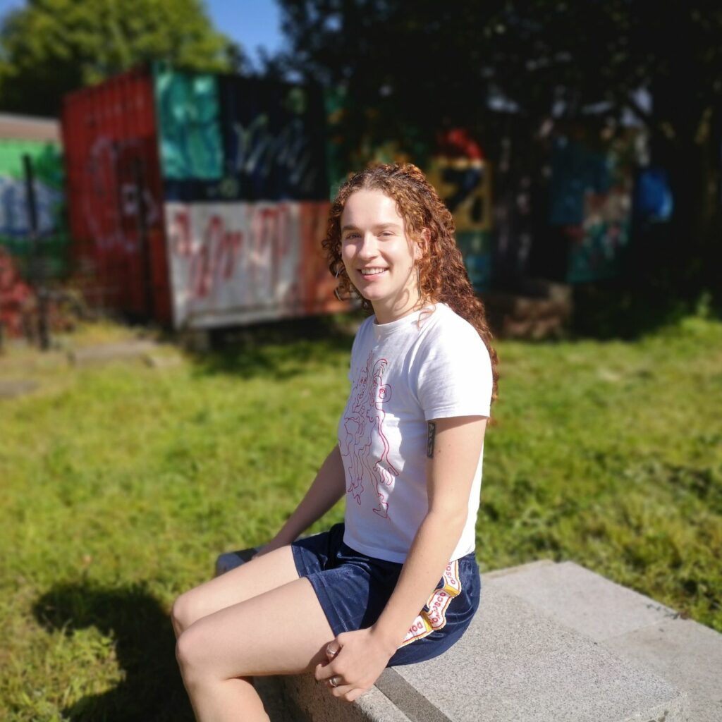 Photo of Hot Desk Member for Ethical Practice Sorcha Perris. Sorcha is sitting outside on a cement block surrounded by green grass. It is sunny and she is in shorts and a t-shirt and is smiling at the camera.