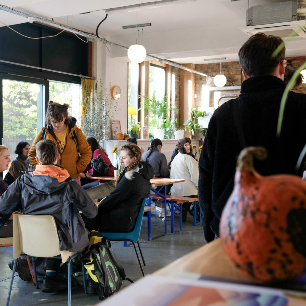 People sit eating and drinking in Civic house canteen where some stalls can be seen in the background. In the very foreground of the photograph is a spherical orange pumpkin.