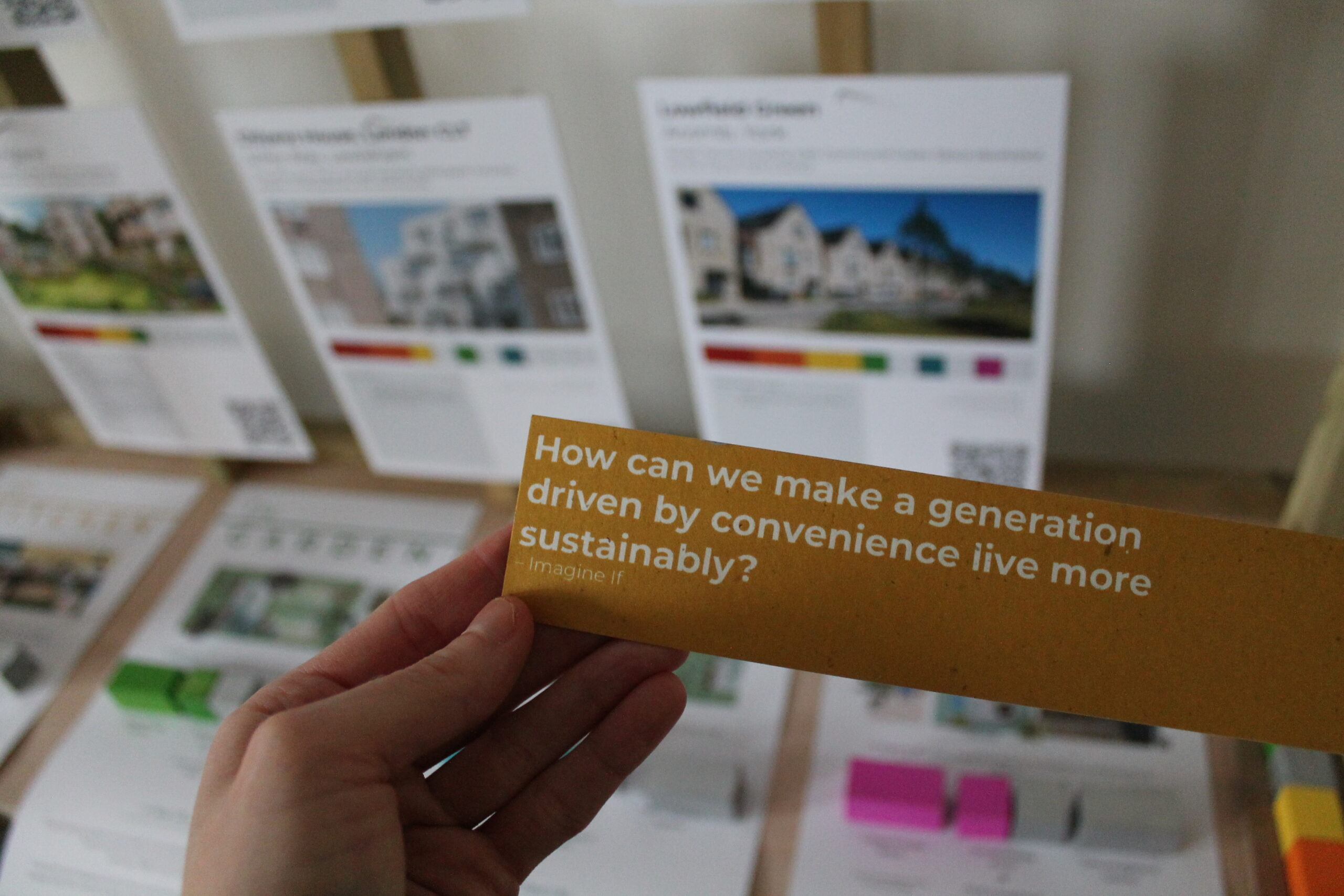 In front of exhibition materials on housing a hand holds up a piece of card which questions how to change a generations impulse of convenience towards sustainable practices.