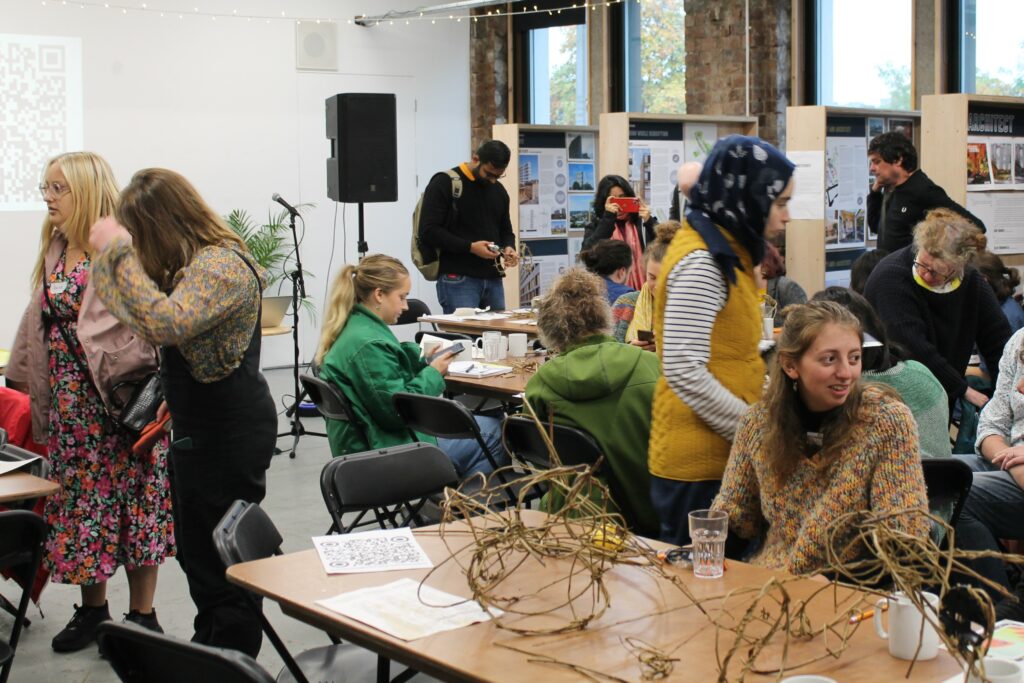 People sit and stand around tables at Civic House Venue, they are engaging in various discussions during a workshop. Behind them are exhibition boards by the windows.