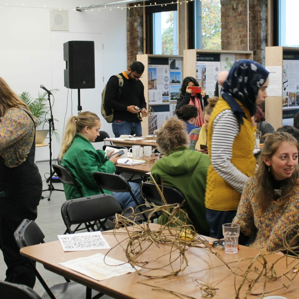 People sit and stand around tables at Civic House Venue, they are engaging in various discussions during a workshop. Behind them are exhibition boards by the windows.