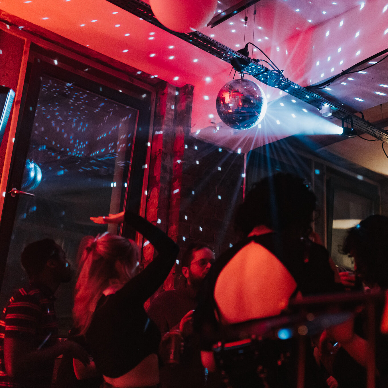 People dance with their backs to us in a room with large windows and a red light. Over their heads is a disco ball lit up by a blue/ white light.